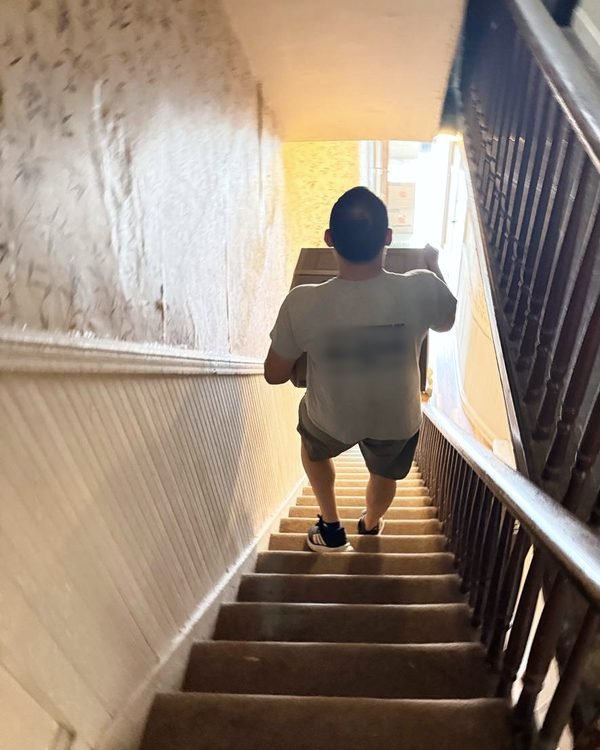 A mover carrying a box up a steep, narrow staircase toward the light