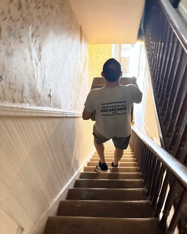 A mover carrying a box up a steep, narrow staircase toward the light